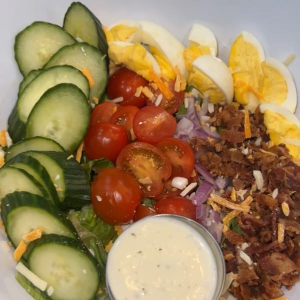 A breakfast salad with cucumbers, cherry tomatoes, hard-boiled eggs, bacon, cheese, and a side of ranch dressing.