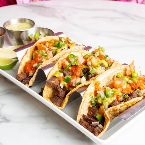 Four breakfast tacos with meat, salsa, and chopped onions on a white platter with lime wedges.