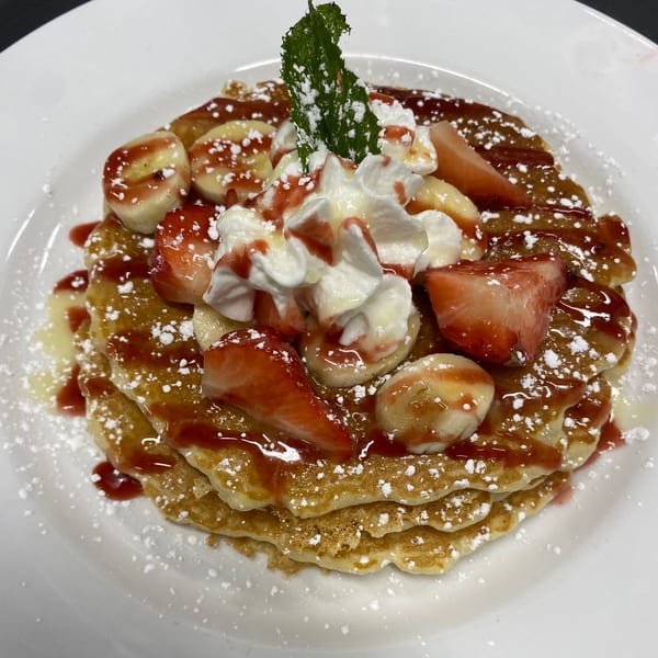 Stack of pancakes topped with strawberries, bananas, whipped cream, strawberry syrup, and powdered sugar.