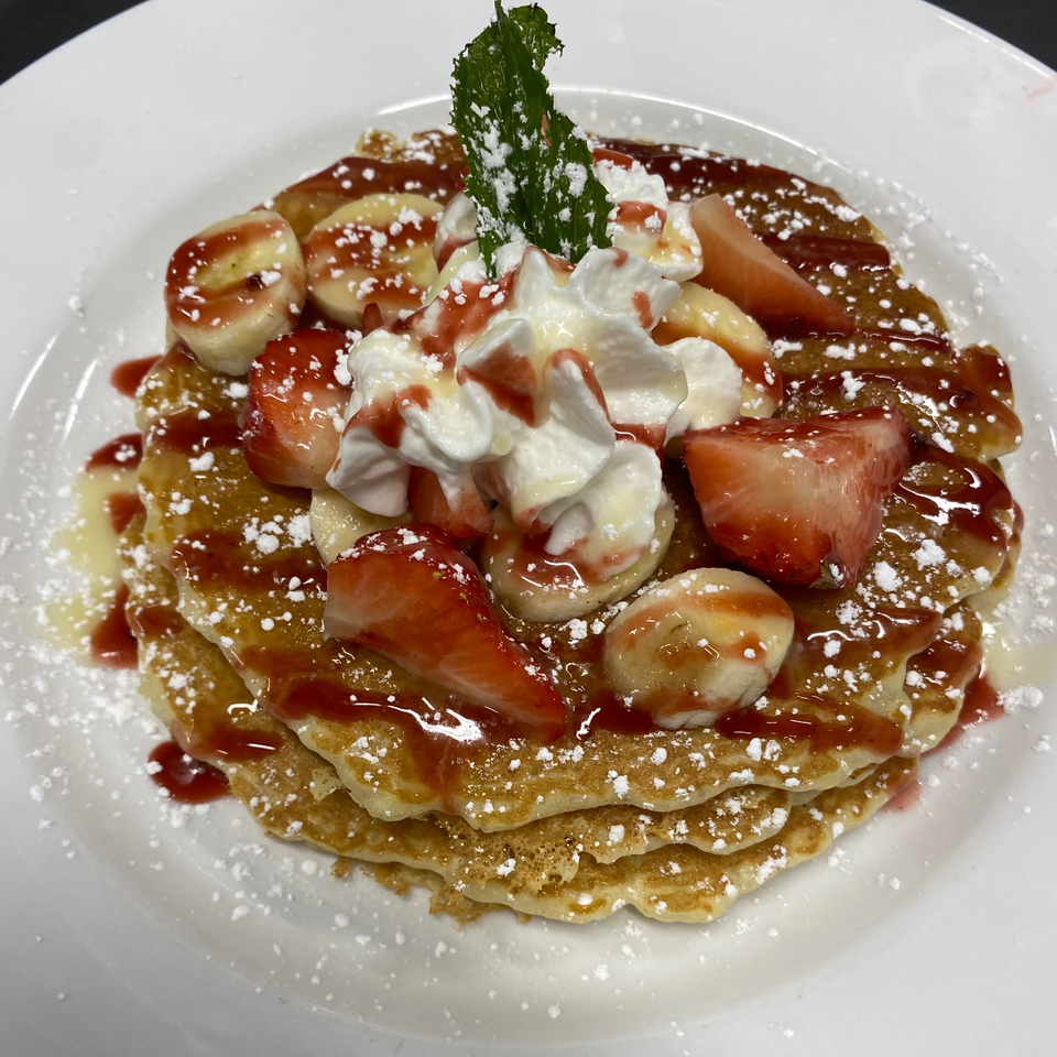 Stack of pancakes topped with strawberries, bananas, whipped cream, strawberry syrup, and powdered sugar.