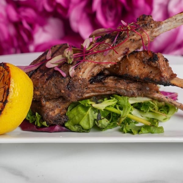 Grilled lamb chops on a bed of greens with a charred lemon half, against a pink floral background.