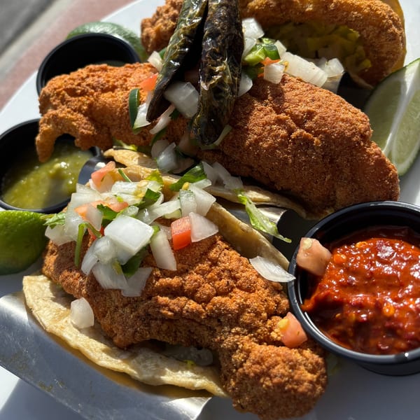 Crispy fried fish tacos topped with pico de gallo, charred peppers, and lime wedges, with salsa and guacamole.