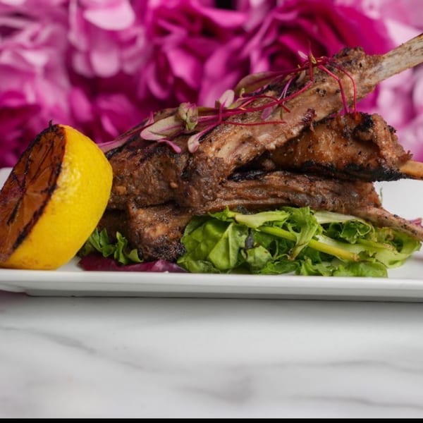 Grilled lamb chops with a side of greens and a charred lemon wedge on a white plate.
