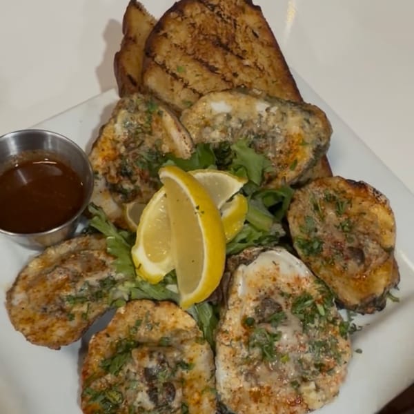 Grilled oysters with lemon wedges, greens, and a side of sauce on a white plate.