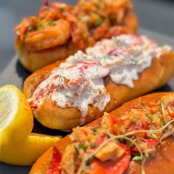 Three lobster rolls on a dark plate, two with creamy white sauce, one with red lobster meat.