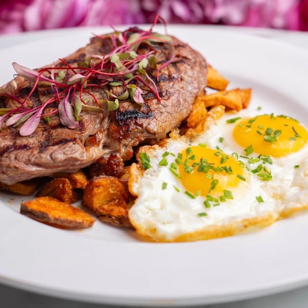 Grilled steak topped with microgreens, served with crispy potatoes and two sunny-side-up eggs with chives.