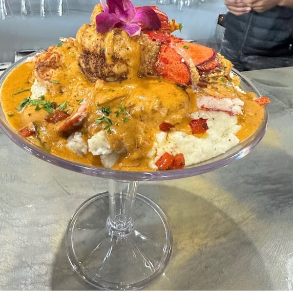 A large glass goblet filled with grits, lobster, and a creamy orange sauce, garnished with a purple orchid.