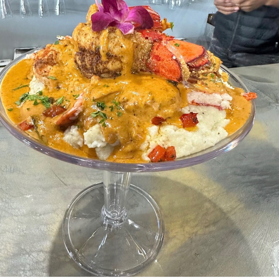 A large glass goblet filled with grits, lobster, and a creamy orange sauce, garnished with a purple orchid.