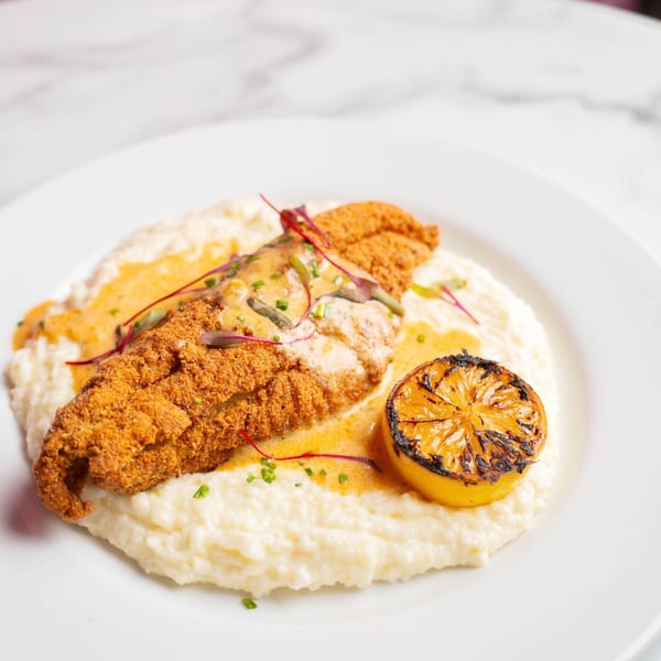 Crispy fried fish fillet with creamy grits, orange sauce, and a grilled lemon on a white plate.