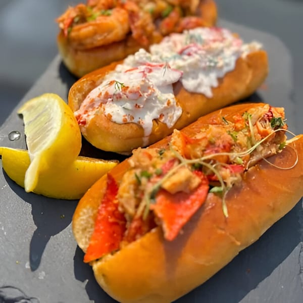 Three lobster rolls, one with red lobster, one with white sauce, and one with shrimp, with lemon wedges.