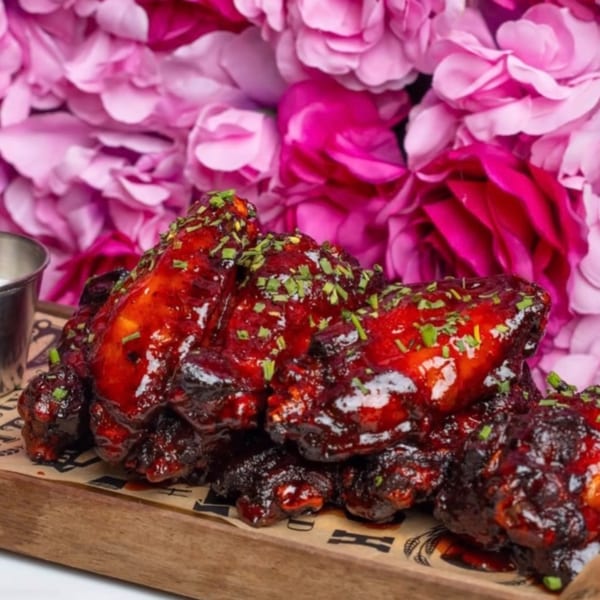 Sweet and spicy chicken wings with chives on a wooden board, against a pink floral background.