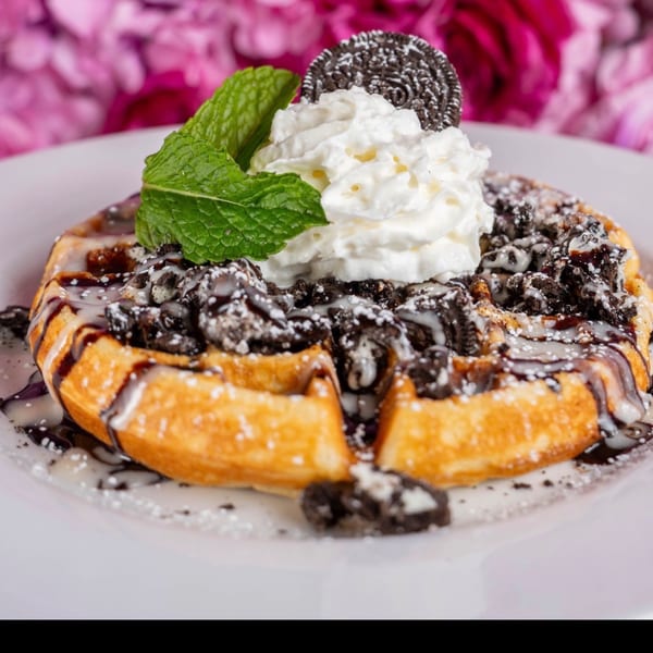 Toast It IZ waffle with whipped cream, Oreo crumbles, chocolate drizzle, and mint, against pink flowers.