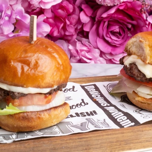 Two mini burgers with cheese, tomato, and lettuce on a wooden tray with pink flowers in the background.
