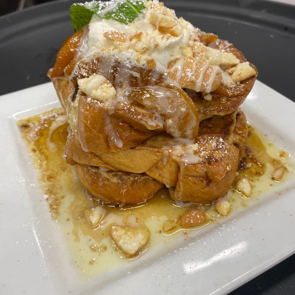 Golden French toast stack, drizzled with syrup and icing, topped with whipped cream, mint, and crumbles.
