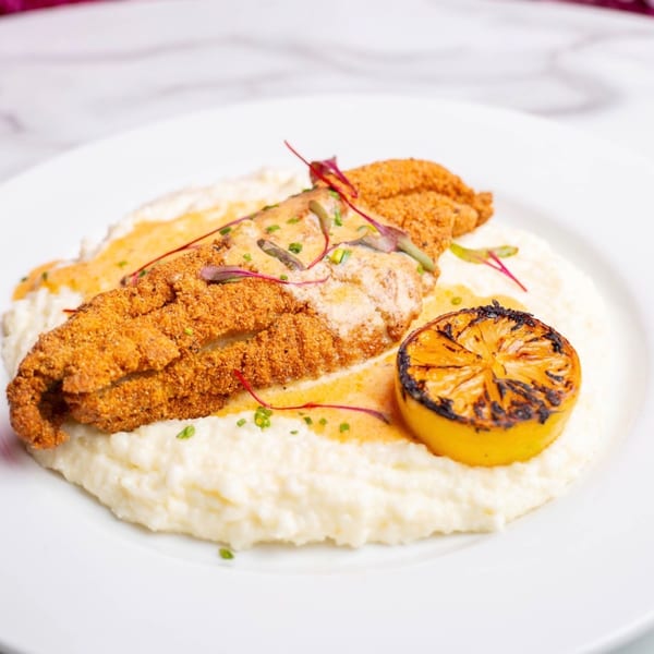 Crispy fried fish fillet on a bed of creamy grits, garnished with sauce, herbs, and a grilled lemon.