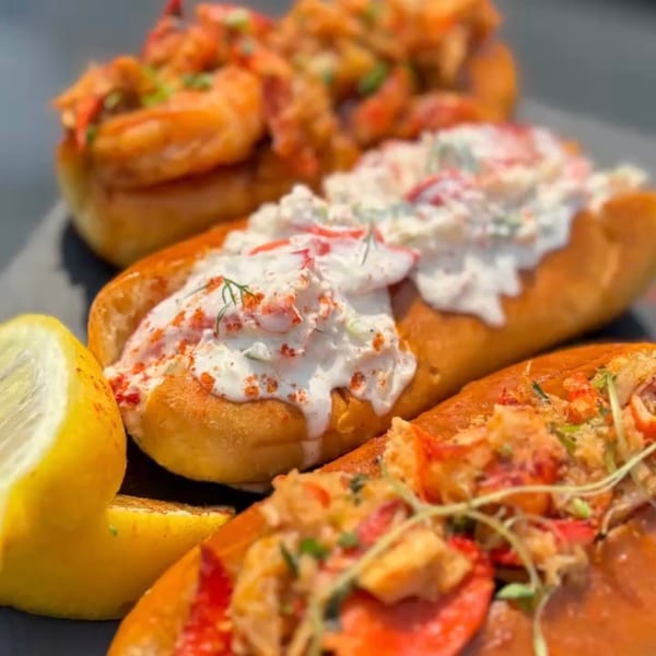 Three lobster rolls with various toppings and a lemon wedge on a dark platter.